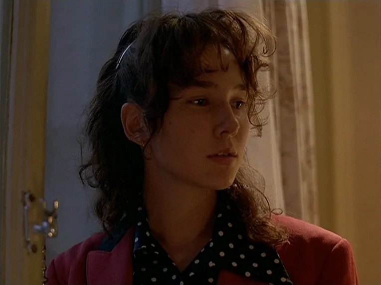 Detective (1985) movie still: closeup — A woman stands in a dim room with curtains visible in the background. She faces slightly away from the…