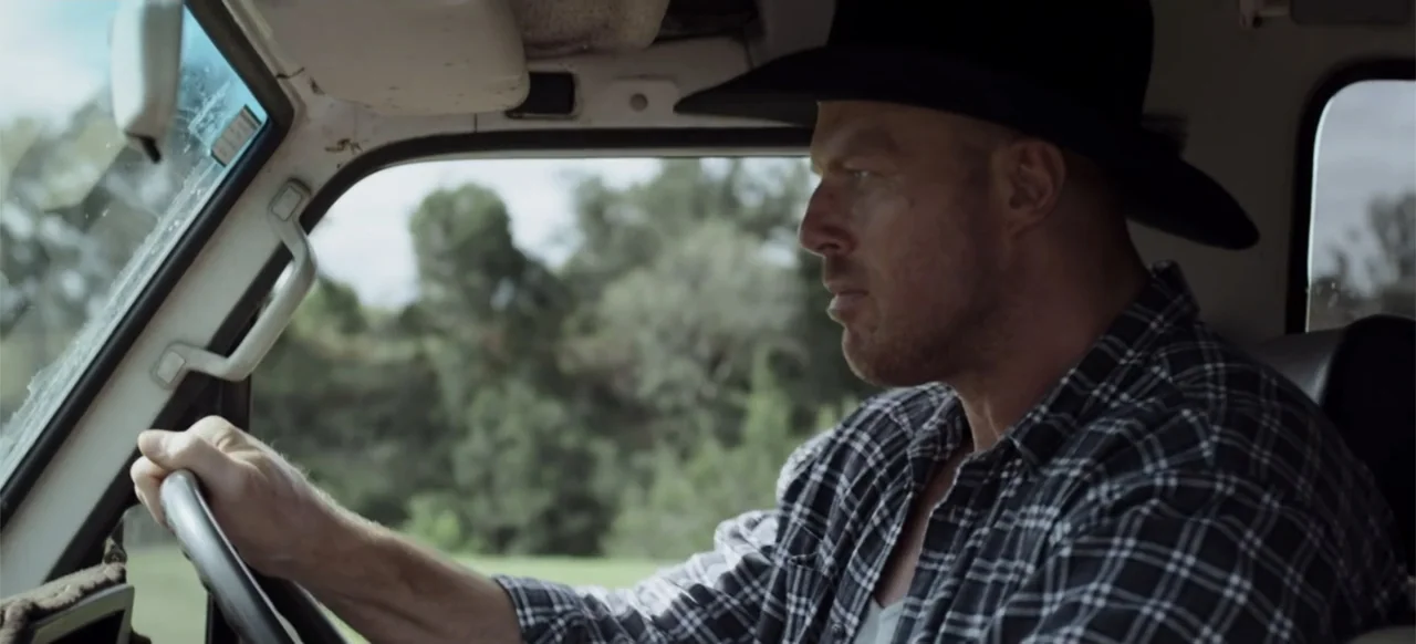 Boar (2018) movie still: medium — The interior of a vehicle shows a man sitting in the driver's seat. A man in a flannel shirt and hat holds the…