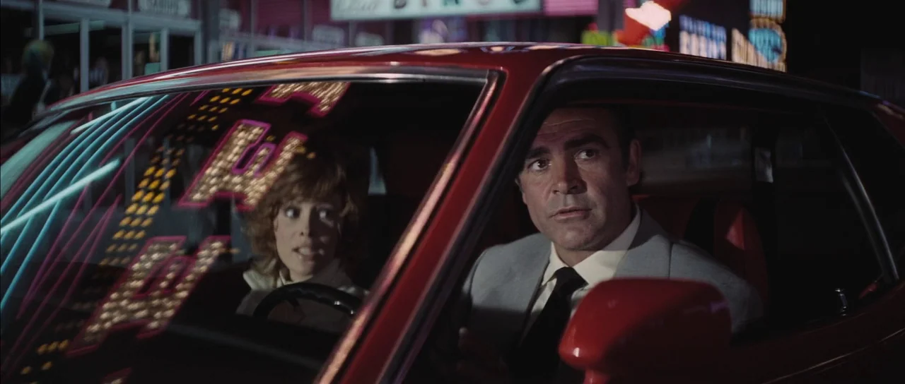 Diamonds Are Forever (1971) movie still: medium — A red vehicle is parked at night in an urban environment with illuminated signs. A man and woman…
