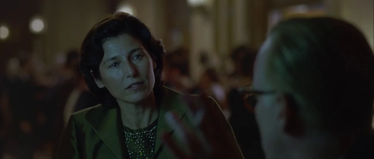 Capote (2005) movie still: medium — A dimly lit indoor hallway with a blurred, crowded background of people. A woman with dark hair speaks while…