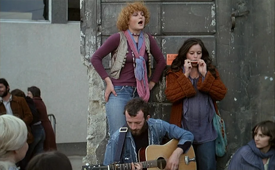 L’une Chante, l’Autre Pas (1977) movie still: fullbody — A group of young musicians stands against a textured concrete wall on a city sidewalk. The…
