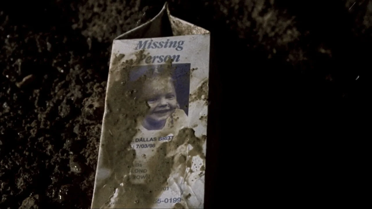 Suspect Zero (2004) movie still: closeup — A milk carton with a missing child flyer lies on the dark wet earth. Mud smears partially obscure the…