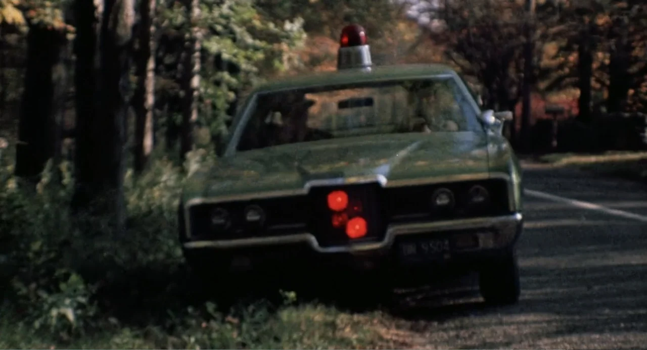 The Last House on the Left (2009) movie still: medium — A dark green vintage sedan is parked on the shoulder of a tree-lined road. The car displays…