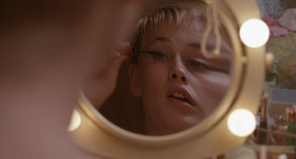 The Indian Runner (1991) movie still: closeup — A woman with short blonde hair gazes into an illuminated vanity mirror. She uses an eyeliner brush…