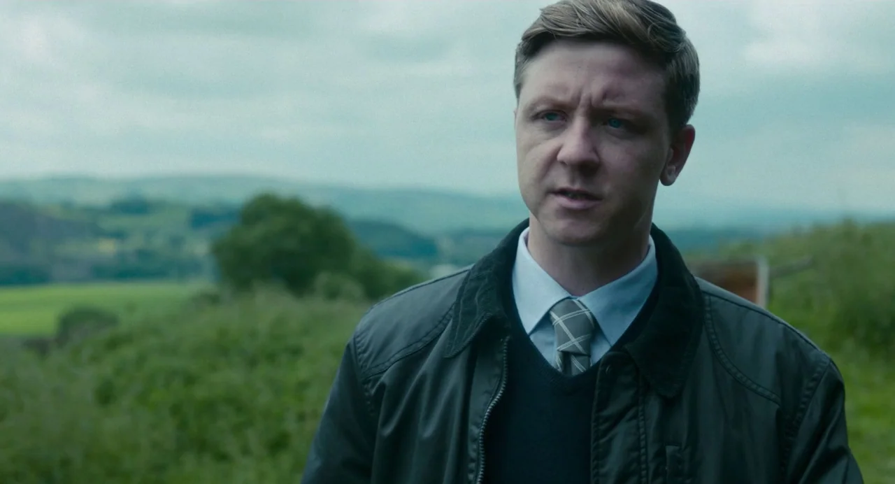 Dark River (2017) movie still: medium — A man stands outdoors in a rolling green field under an overcast sky. The subject is wearing a collared…