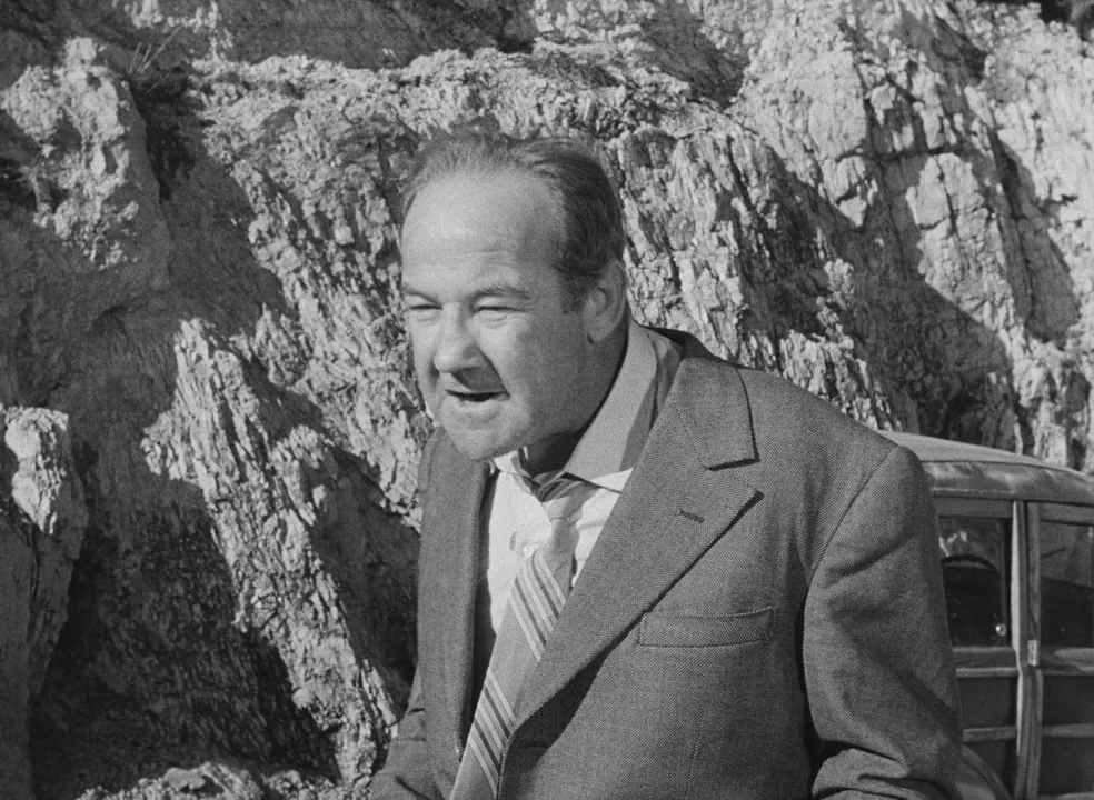 Il Bidone (1955) movie still: medium — A man in a suit stands in front of a jagged limestone cliff face. A car is visible partially on the right…