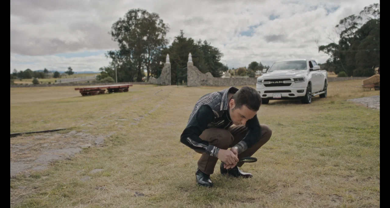 National Family (2023) movie still: medium — An open field contains a white pickup truck and a stone gateway in the distance. A man crouches in the…