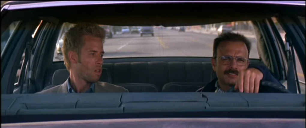 Memento (2000) movie still: medium — The interior of a moving sedan shows a city street visible through the windshield. Two men sit in the front…