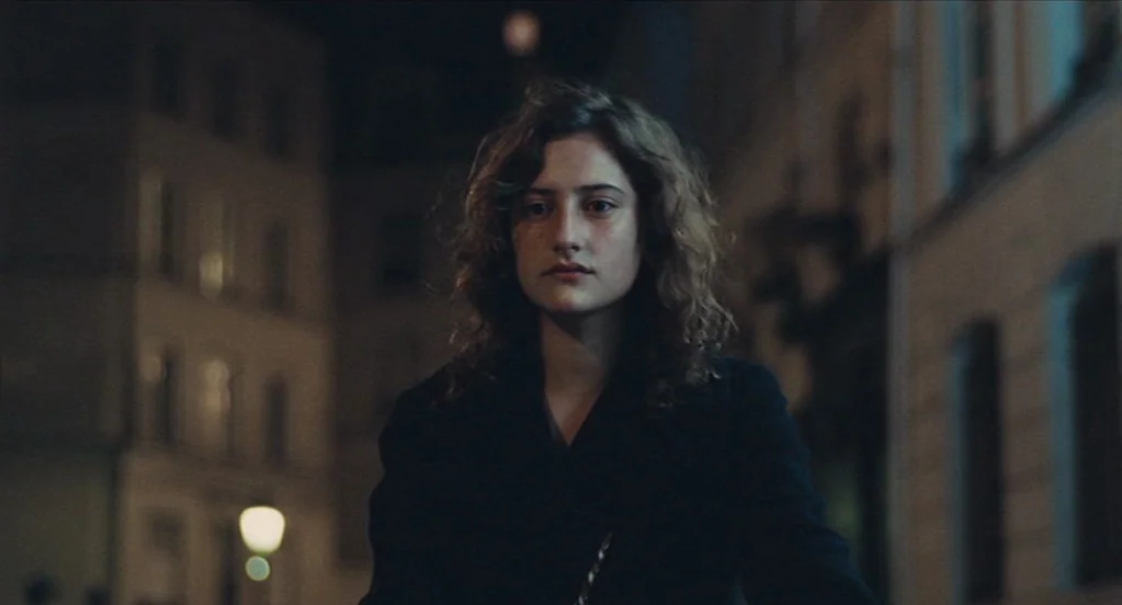 Something in the Air (2012) movie still: medium — A woman stands in the middle of a dimly lit city street at night. Tall buildings blur into…
