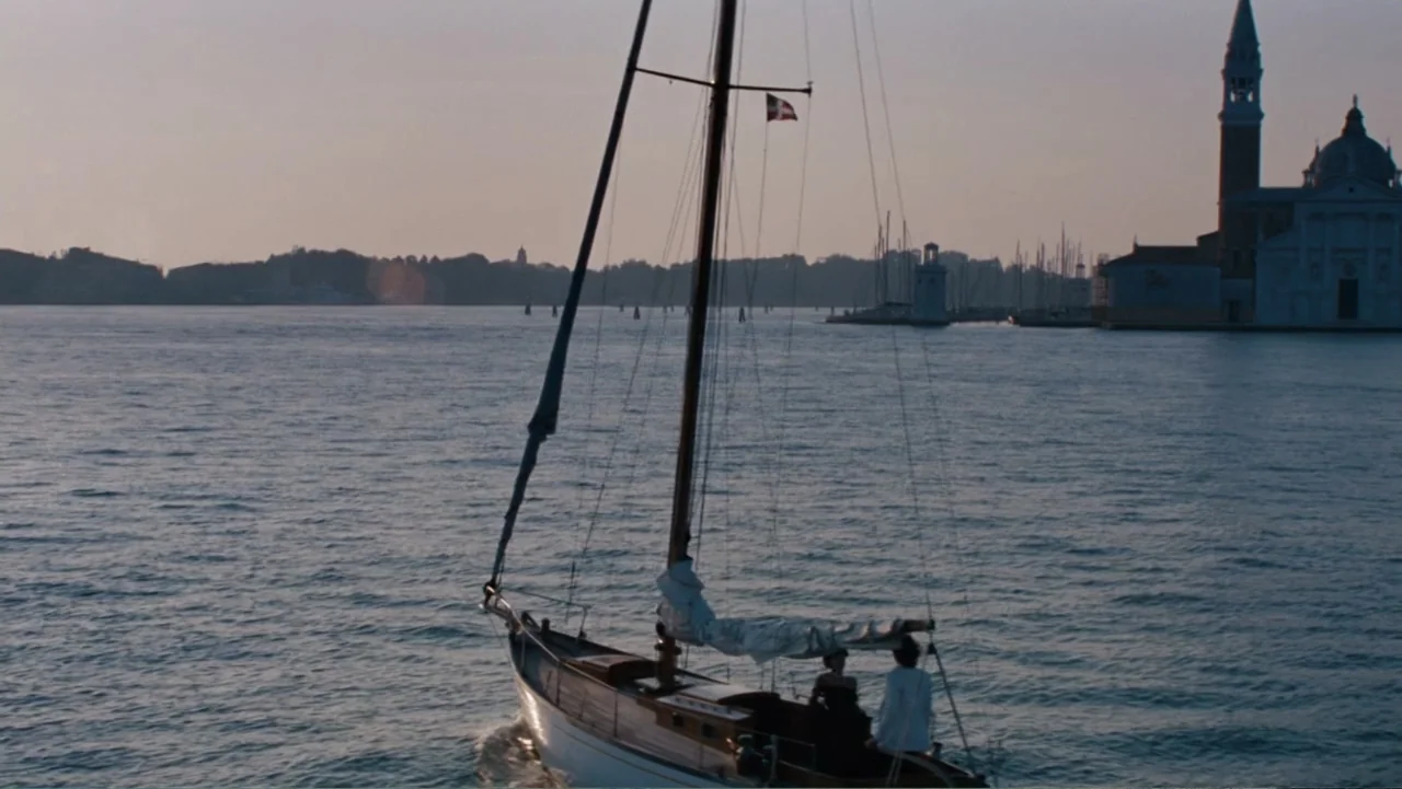 The Tourist (2010) movie still: wide — A wooden sailboat moves across a Venetian lagoon with historic church architecture visible behind. Two…
