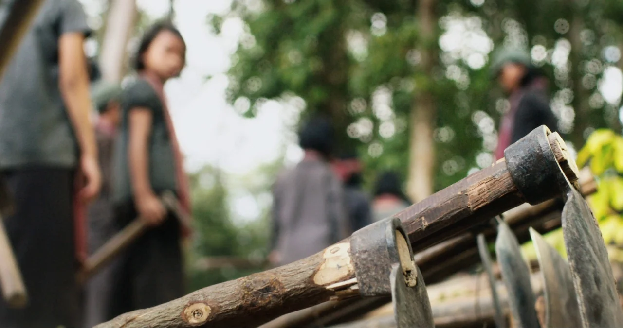 First They Killed My Father (2017) movie still: closeup — A collection of agricultural hoes with wooden handles sit in the foreground of a forest.…