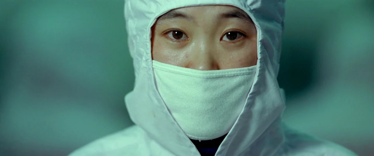 Samsara (2011) movie still: closeup — A person in a sterile white protective suit stands within a clinical laboratory environment. The subject looks…