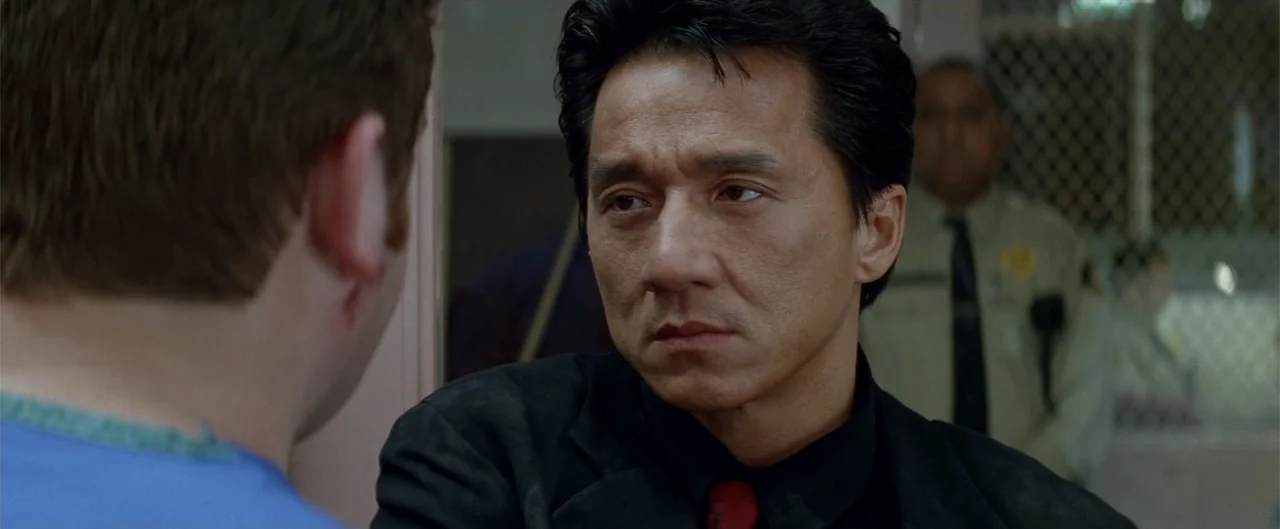 Rush Hour (1998) movie still: medium — An interview room has a metal mesh divider visible behind a seated man. A man in a dark suit talks to a…