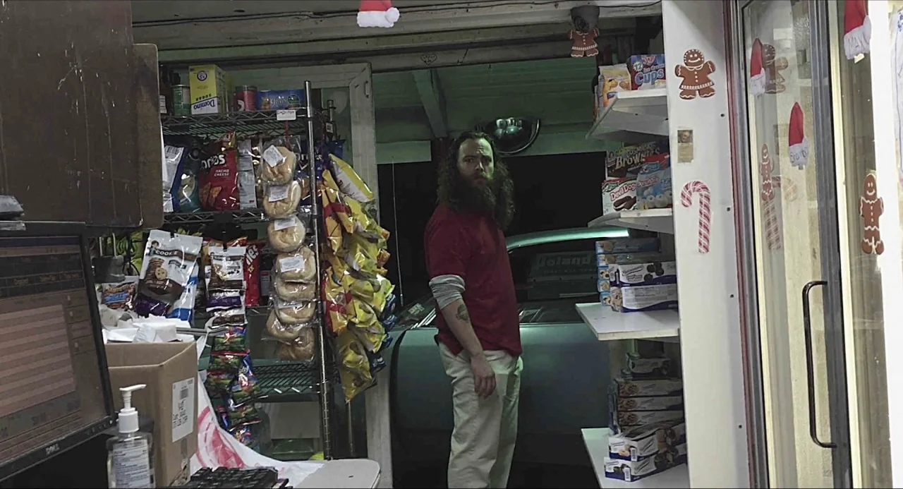 Like Me movie still: medium — A cluttered convenience store checkout area features metal shelving filled with bagged snacks. A bearded man in a…