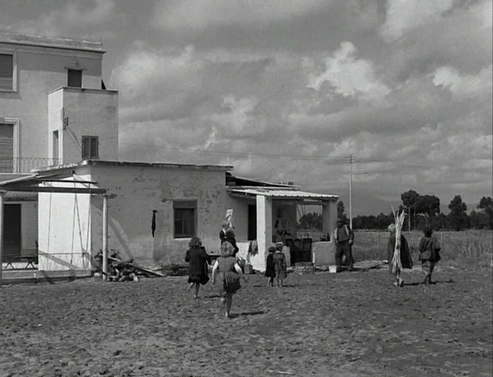 La Strada (1954) movie still: wide — A low white building stands on a dusty, barren landscape under a cloudy sky. Several children are running…
