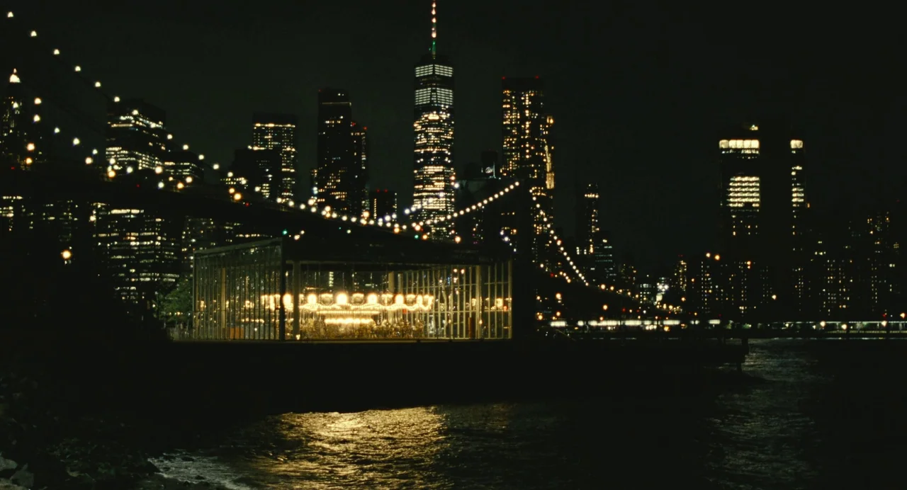 Past Lives (2023) movie still: establishing — A glass-enclosed carousel sits on the Brooklyn waterfront at night under the bridge lights. The…
