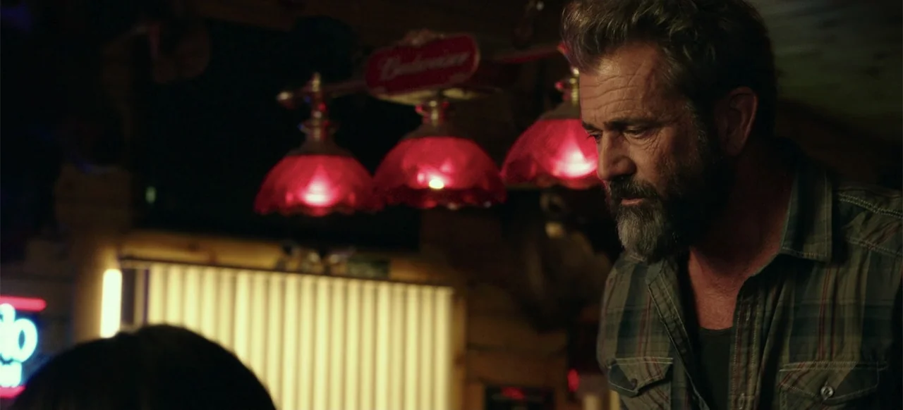 Blood Father (2016) movie still: medium — A dimly lit bar setting with red glass lamp fixtures hanging in the background. An older man with a beard…