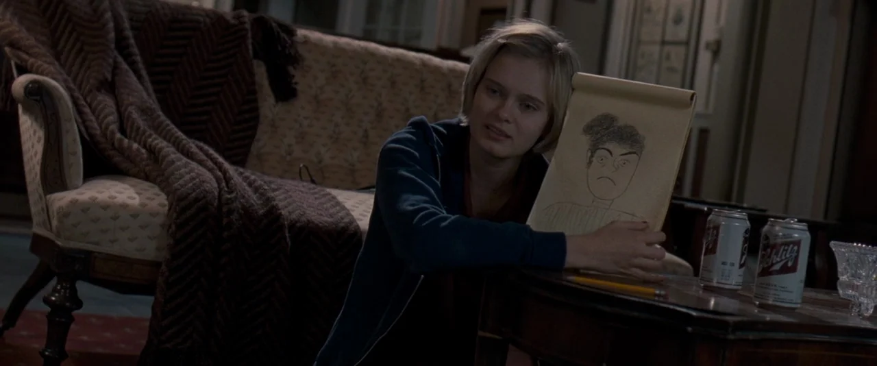 The Innkeepers (2011) movie still: medium — A dark, dimly lit room contains an upholstered chair and a small side table. A young woman rests her arm…