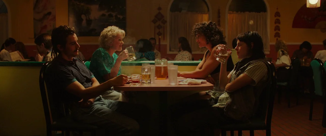 Love Lies Bleeding (2024) movie still: wide — A retro restaurant booth is dimly lit with amber overhead light and tiled flooring. Four adults sit…