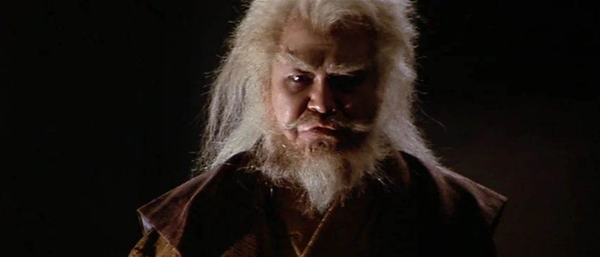 Lone Wolf and Cub: Baby Cart in Peril (1972) movie still: closeup — An older man with long white hair stands against a pitch black background. He…