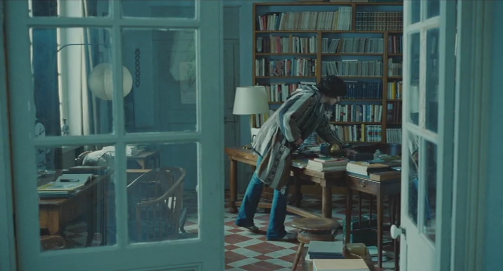 Something in the Air (2012) movie still: medium — A room with blue walls features a floor covered in white and red tiles. A man stands at a wooden…