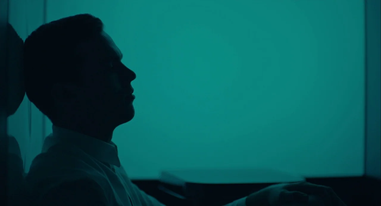 Equals (2015) movie still: medium — A man sits in a stark, dimly lit room illuminated by a large screen. He rests his head against the wall while…