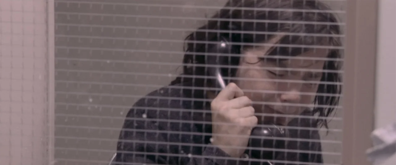 Dancer in the Dark (2000) movie still: closeup — A person sits behind a security grid in a nondescript, light-colored room. The person holds a black…