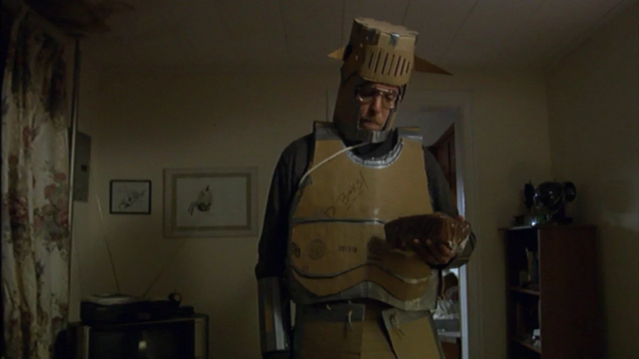 Murder Party movie still: medium — A dimly lit room with floral curtains and framed pictures on plain walls. A man wearing elaborate cardboard armor…