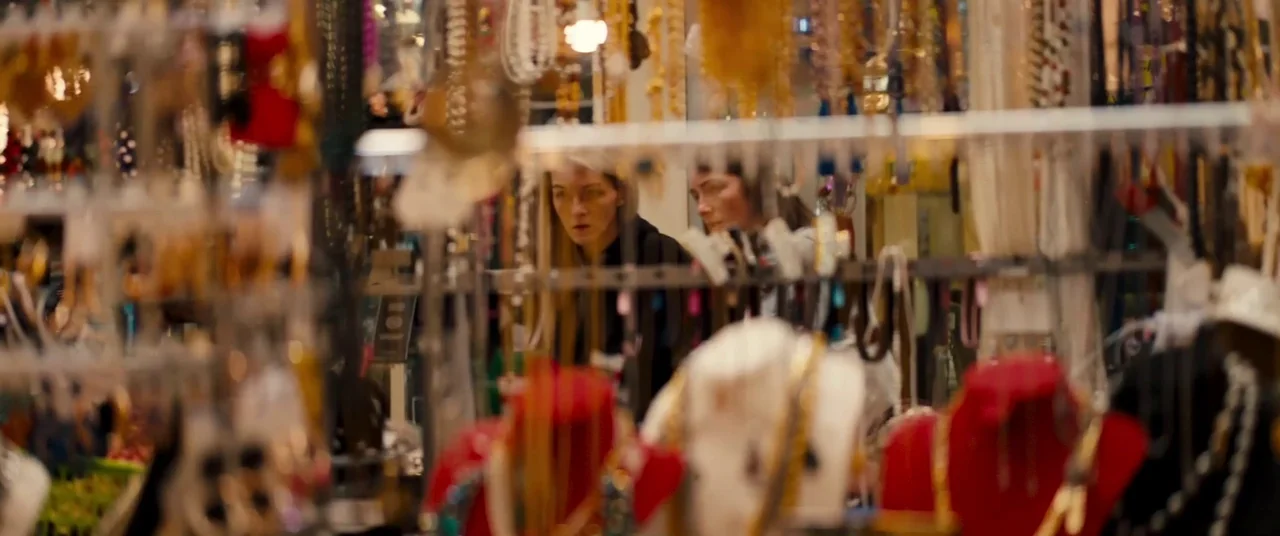 Silver Haze (2023) movie still: medium — A dense display rack of necklaces and jewelry fills the foreground of a shop. Two women stand behind the…
