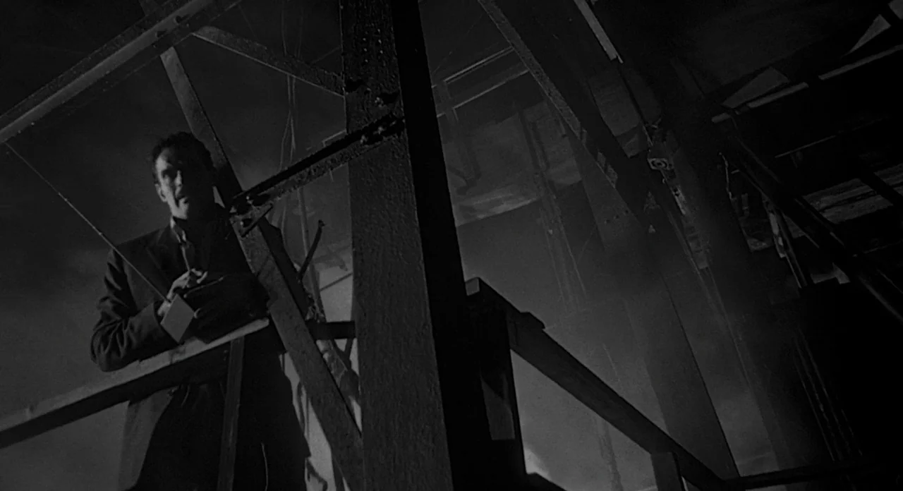Touch of Evil (1958) movie still: medium — An industrial platform features crisscrossing steel beams and support struts in a dark space. A man…