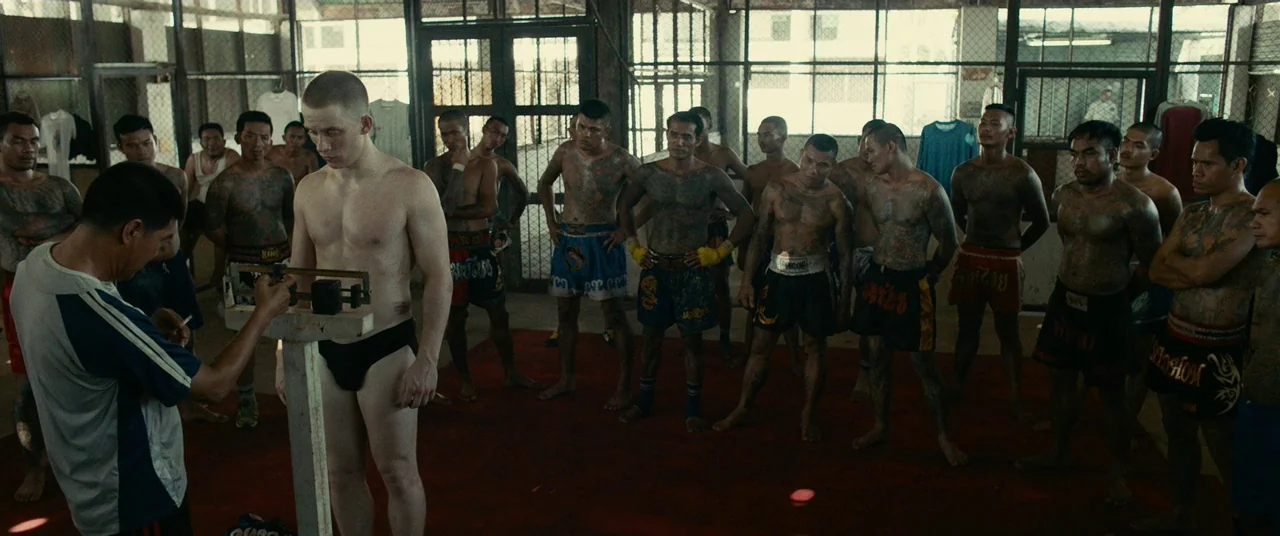 A Prayer Before Dawn (2017) movie still: fullbody — A group of shirtless men with various tattoos stands inside a Thai boxing gym. A young man…