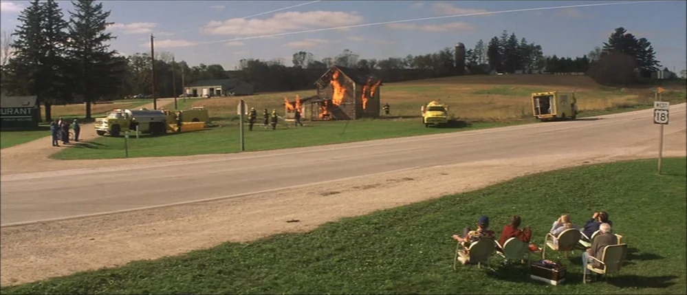 The Straight Story (1999) movie still: wide — A rural landscape features a burning wooden house beside a quiet road. A group of onlookers sit in…