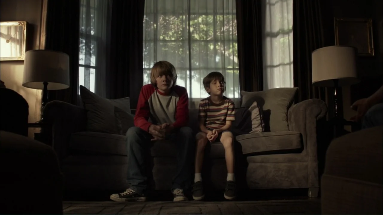 Dark Skies (2013) movie still: medium — A traditional living room features a grey couch positioned in front of a window. Two young boys sit together…