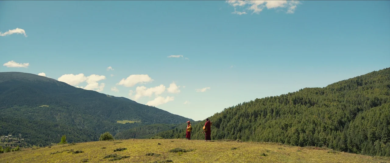 The Monk and The Gun (2023) movie still: wide — A high, grassy mountain ridge overlooks a vast valley covered in dense pine forest. Two figures in…