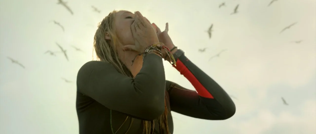 The Shallows (2016) movie still: medium — A woman in a wetsuit is positioned in open water against a pale sky. Multiple gulls fly in the background…