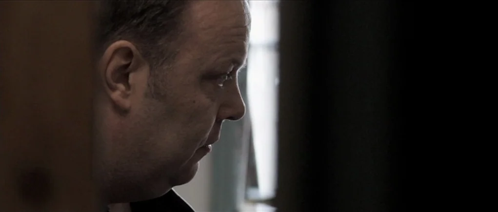 Down Terrace (2009) movie still: closeup — A dark indoor space with a sliver of bright window light in the background. A man stands in profile…