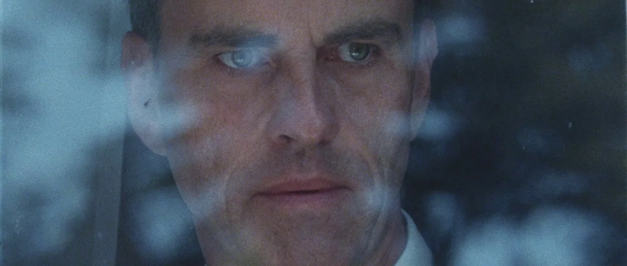 Alléluia (2014) movie still: closeup — A man's face is partially obscured by reflections on a pane of glass. He looks ahead with a steady expression…