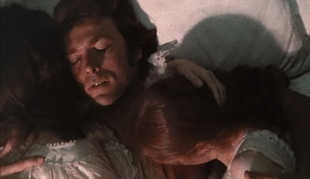 The Beguiled (1971) movie still: closeup — A man lies between two women in a bed with off-white linens. They are all asleep in a dimly lit,…