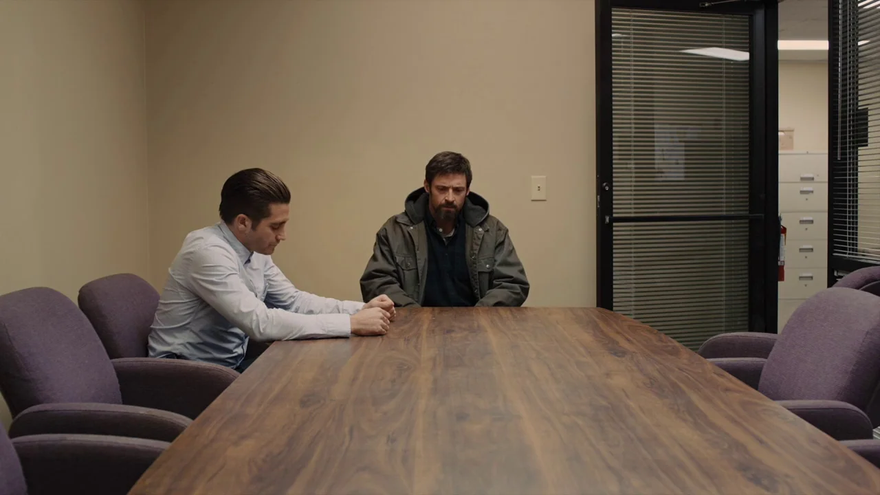 Prisoners (2013) movie still: medium — A spare conference room features plain beige walls and purple upholstered chairs around a table. Two men sit…
