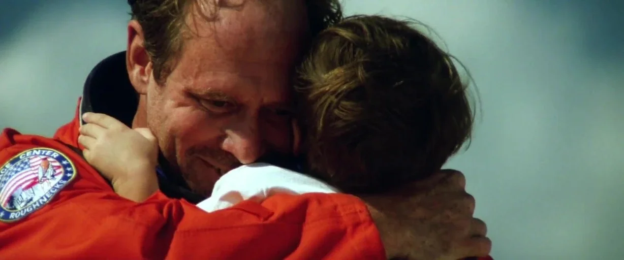 Armageddon (1998) movie still: closeup — A man wearing a bright orange flight suit hugs a child outdoors. The bright sky provides a simple, neutral…