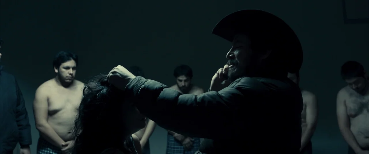 Frontera (2014) movie still: medium — A dimly lit, bare industrial space serves as the backdrop for the gathering. A man in a cowboy hat stands…
