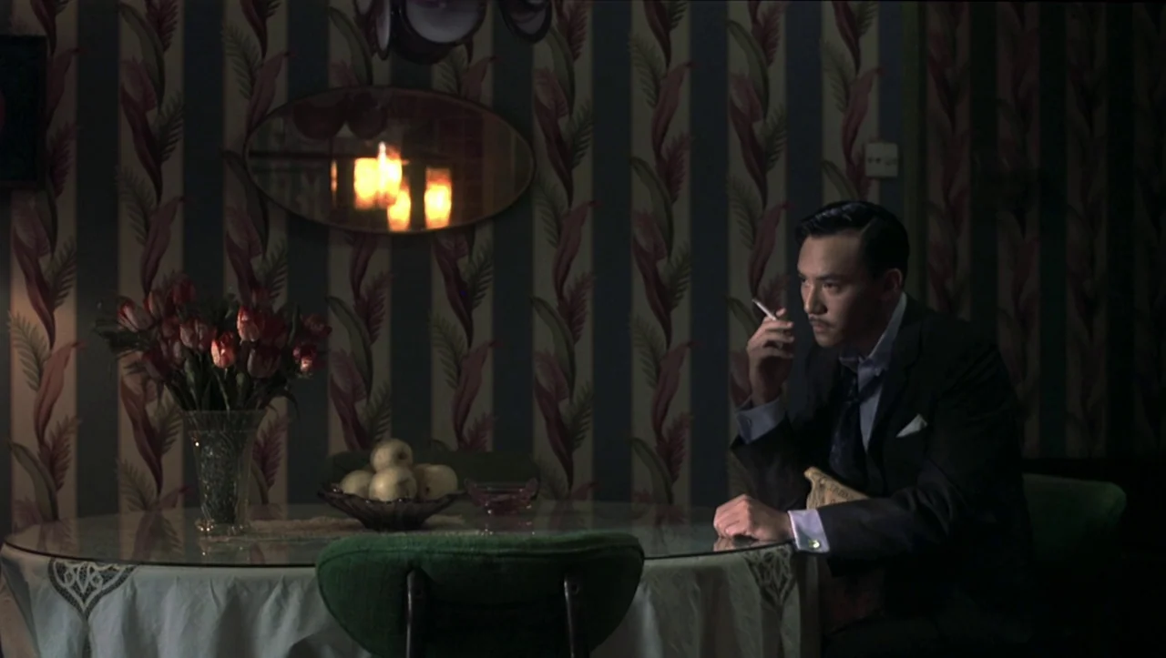 Eros (2004) movie still: medium — A dark dining room features patterned wallpaper and a glass table with a vase. A man in a suit sits at the table…