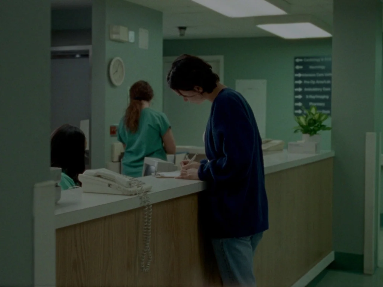 Mid90s (2018) movie still: medium — A medical facility reception area features pale green walls and a laminate counter. A woman in a blue sweater…
