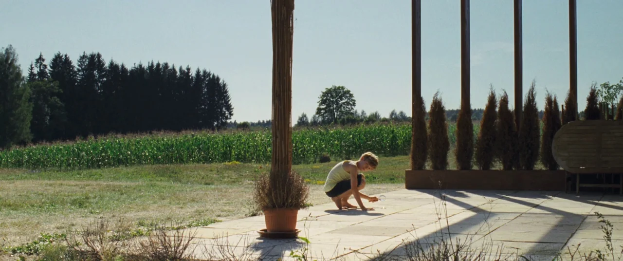 Goodnight Mommy (2014) movie still: wide — A sunny stone patio overlooks a vibrant green cornfield backed by a dense forest. A young child crouches…