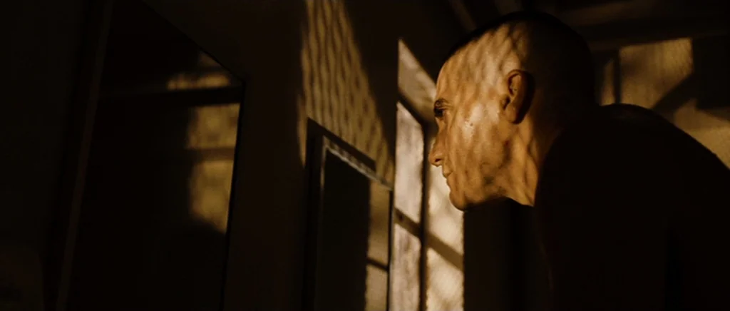 Jarhead movie still: closeup — A dark space features a window with a metal security grate. A man with a buzz cut gazes out through the bars at the…