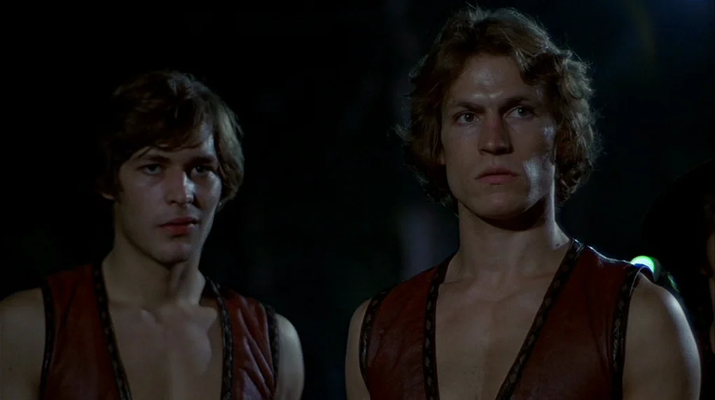 The Warriors (1979) movie still: medium — Two men stand close together in the darkness against an indistinct black background. Both subjects face…