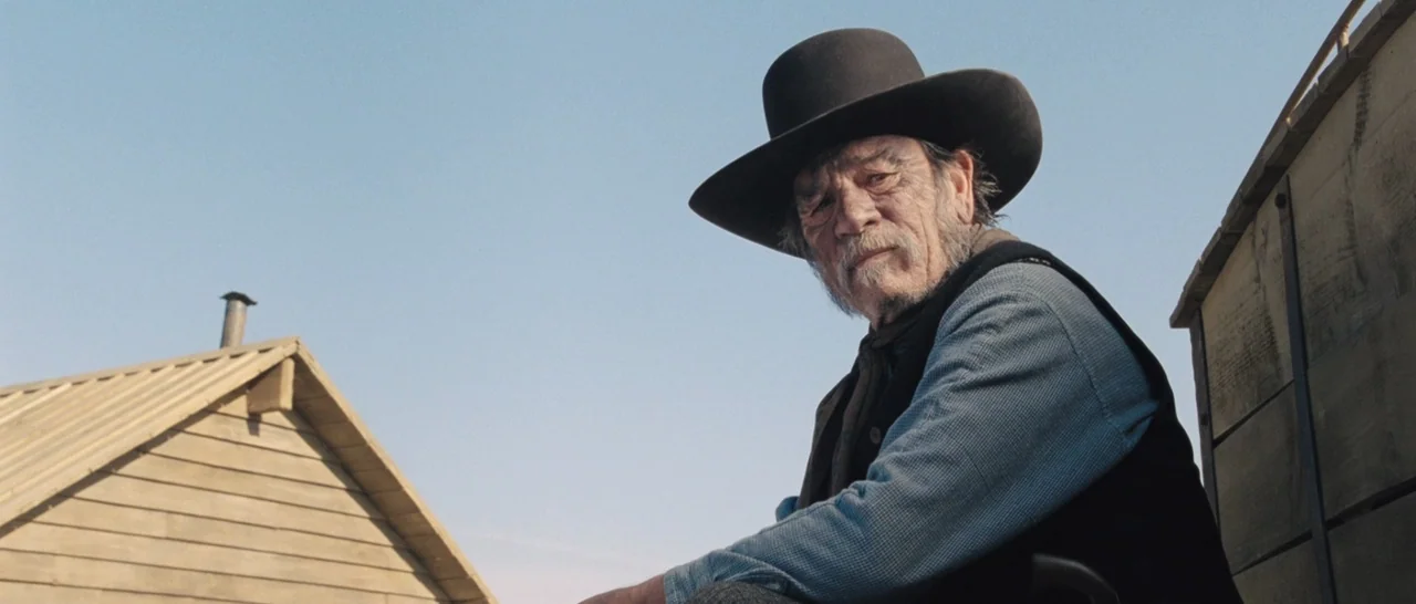 The Homesman (2014) movie still: medium — An elderly man sits on the side of a wooden cargo wagon outdoors. The clear blue sky and the sloped wooden…