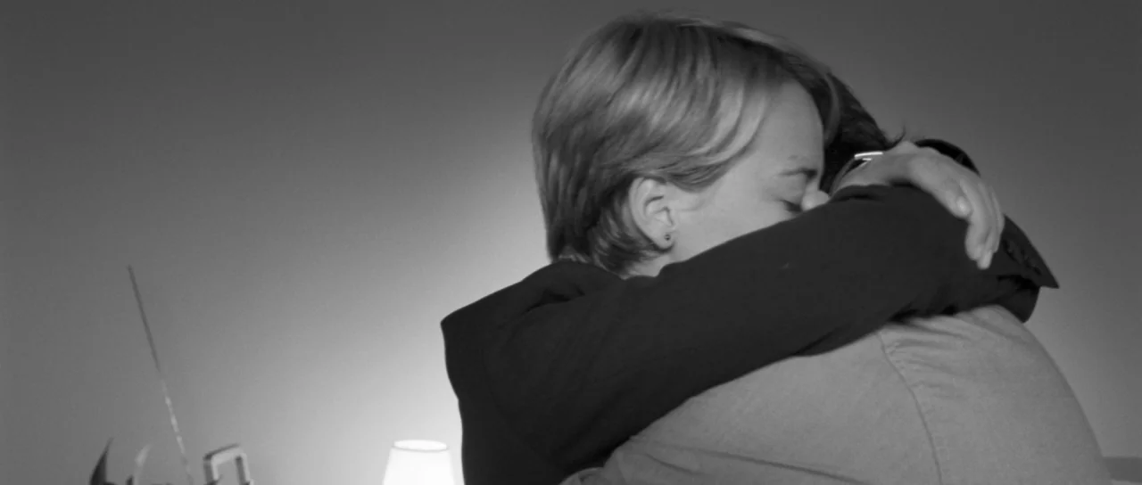 Polytechnique (2009) movie still: closeup — Two individuals embrace closely in a dark interior space with minimal furniture. The woman's face is…