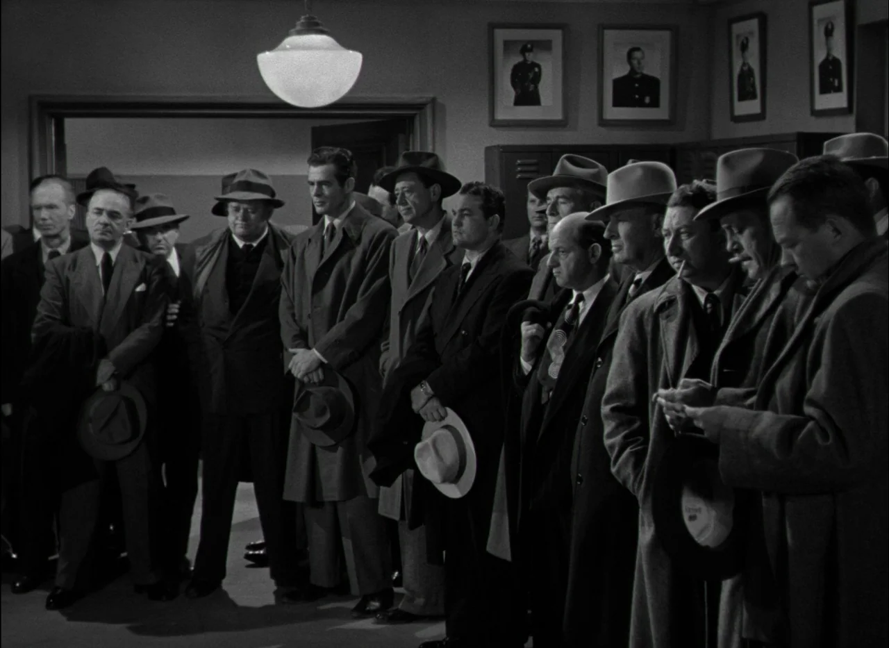 On Dangerous Ground (1951) movie still: medium — A group of men in suits and fedoras stand in a row inside a building. They hold their hats at their…