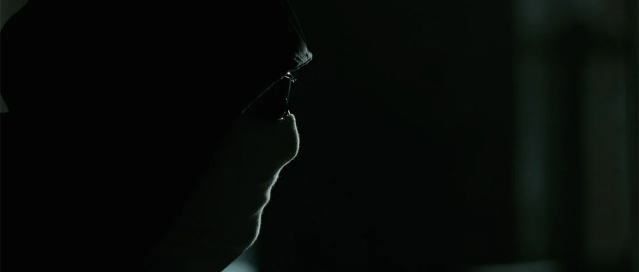 Inside Man (2006) movie still: closeup — A person wears a dark hood that obscures the majority of their facial features. The subject is captured in…